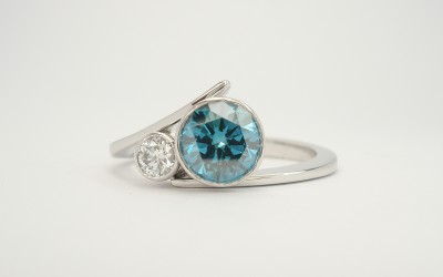 Ocean Blue 1.00ct round brilliant cut diamond & smaller white diamond rub-over set (bezel set) wishbone cross-over style platinum ring.