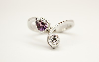 Pear shaped purple sapphire and round brilliant cut diamond rub-over set 2 stone curly wishbone style ring mounted in palladium & platinum.
