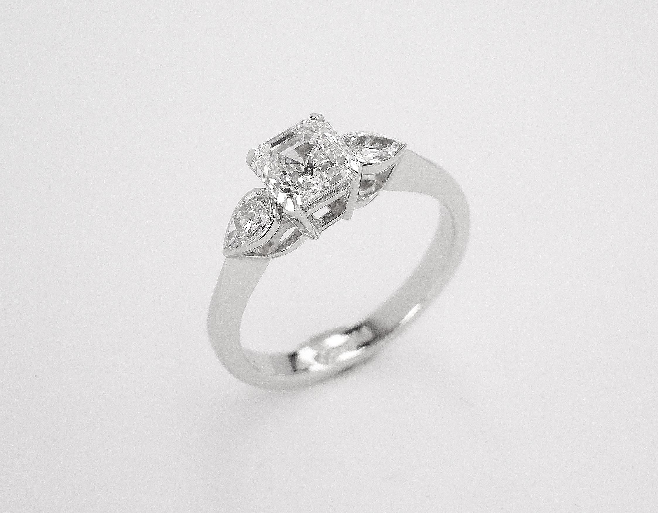 A single 0.82ct. 'E' colour, VS clarity Asscher cut diamond accompanied by a rub-over set pear shaped diamond on each shoulder of the platinum.
