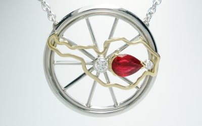 A pear shaped ruby and round brilliant cut 3 stone palladium mountain bike wheel style pendant with 18ct. yellow gold Austrian map overlay.