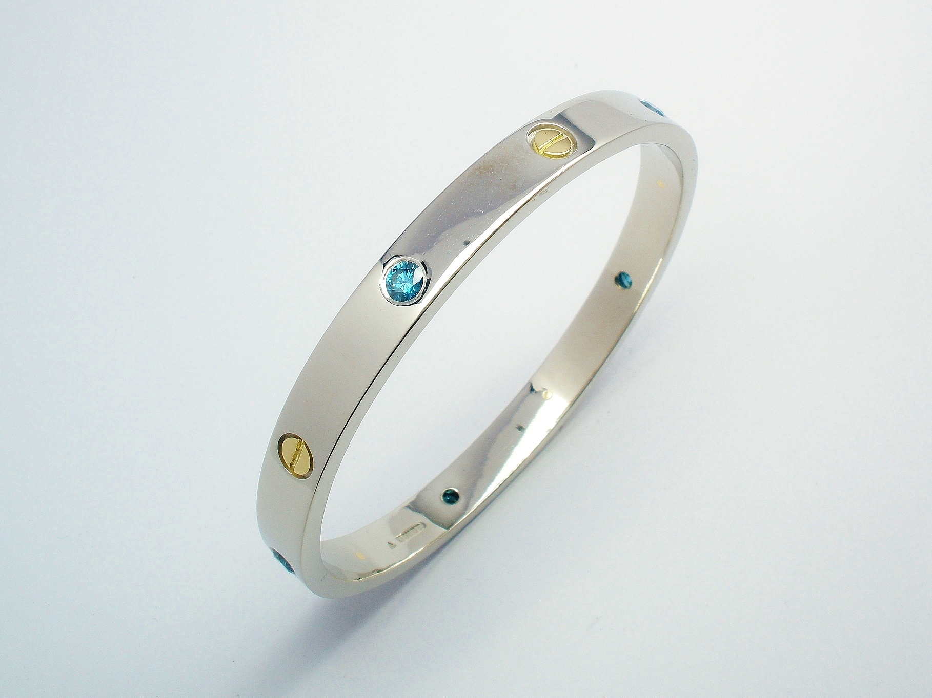 A solid palladium bangle with 18ct. yellow gold 'screw heads' inlayed and flush set with ocean blue round brilliant cut diamonds evenly spaced around and alternating with the screw heads.