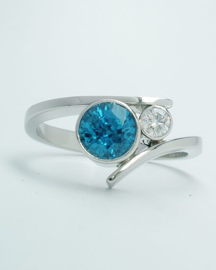 A platinum 2 stone rub-over set round blue Zircon and brilliant cut diamond straight wishbone cross-over dress ring.