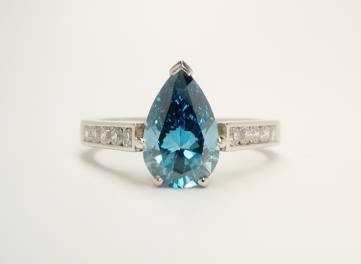 A single stone HTHP treated ocean blue pear shaped diamond ring mounted in platinum with channel set white round brilliant cut diamond shoulders.