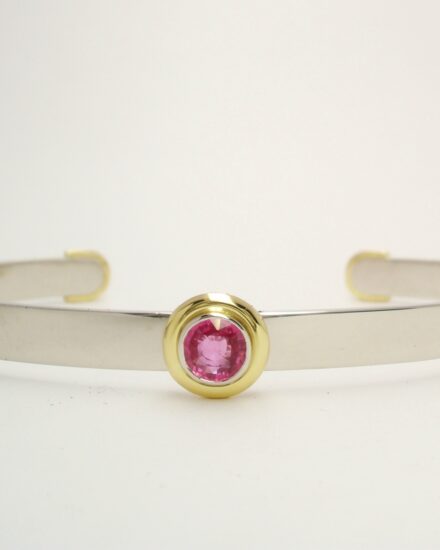 A single stone round pink sapphire torque bangle mounted in platinum and 18ct. yellow gold. The sapphire is rub-over set in platinum with an 18ct. yellow gold 'doughnut' surround.
