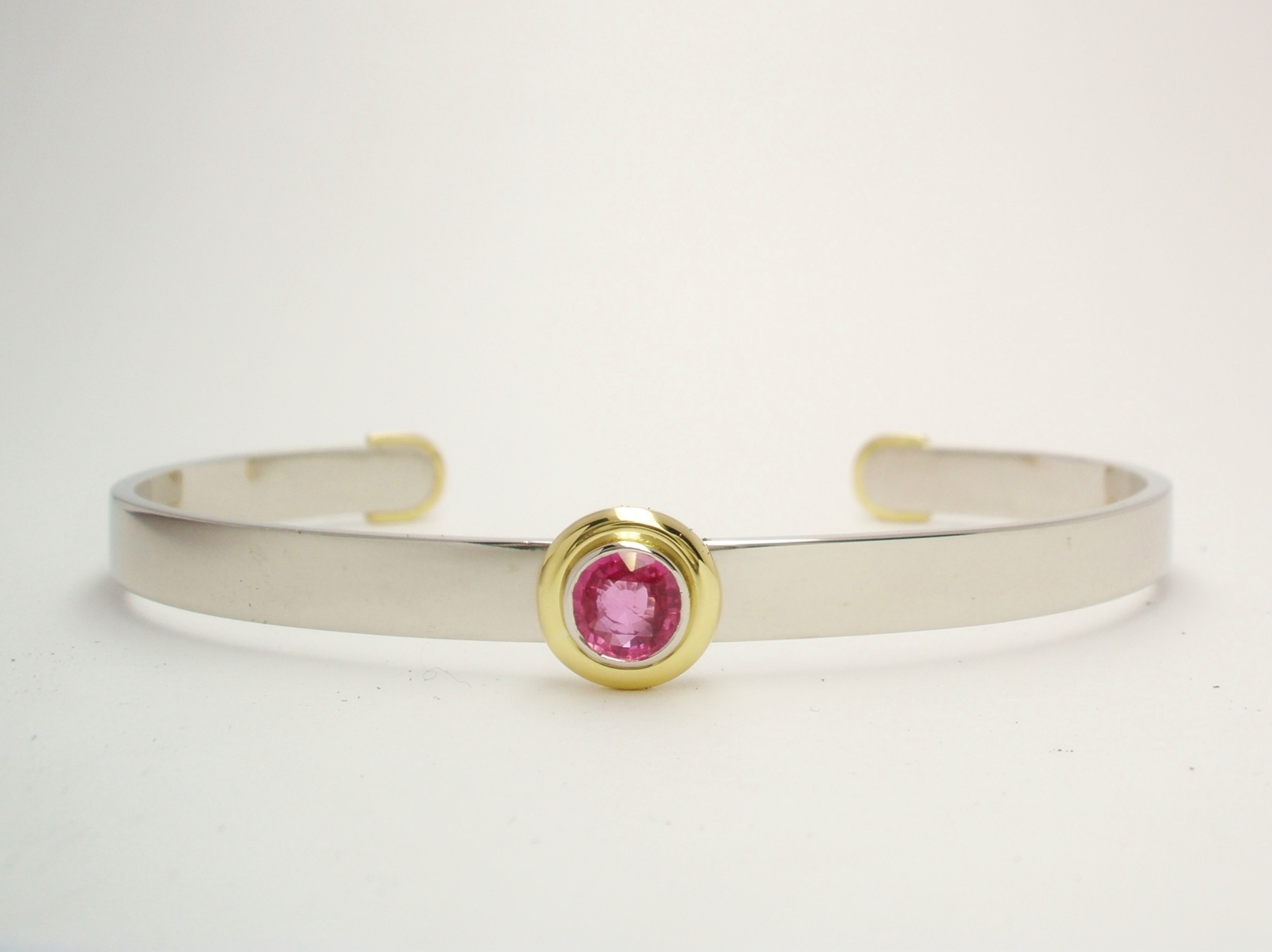A single stone round pink sapphire torque bangle mounted in platinum and 18ct. yellow gold. The sapphire is rub-over set in platinum with an 18ct. yellow gold 'doughnut' surround.