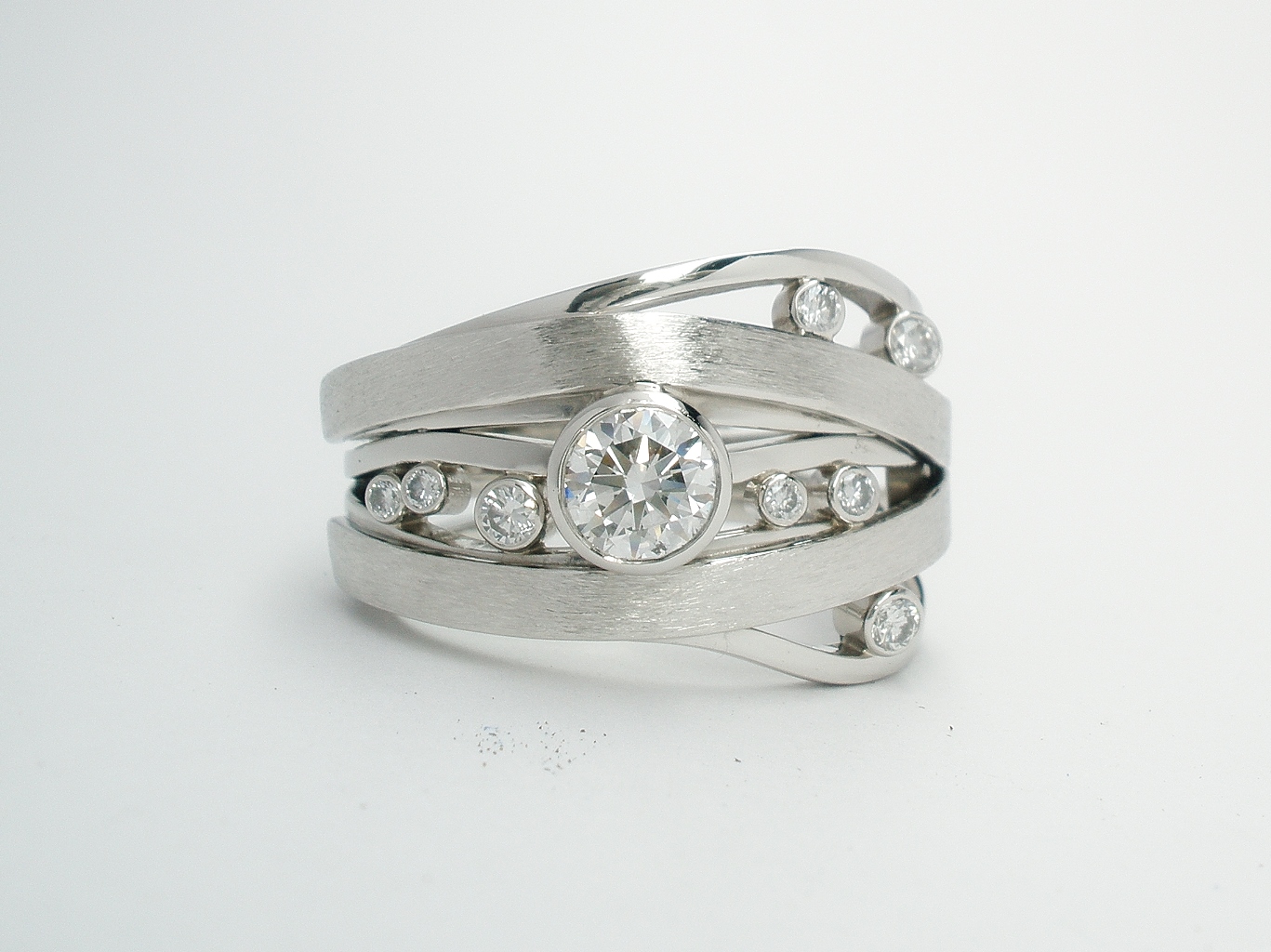 A 9 stone round brilliant cut diamond ring mounted in platinum. The ring consists of five 1mm broad bands that meander around with 2 straps overlayed. The main centre diamond is rub-over set in a raised setting with the 8 smaller diamonds rub-over set low between the 1mm bands.