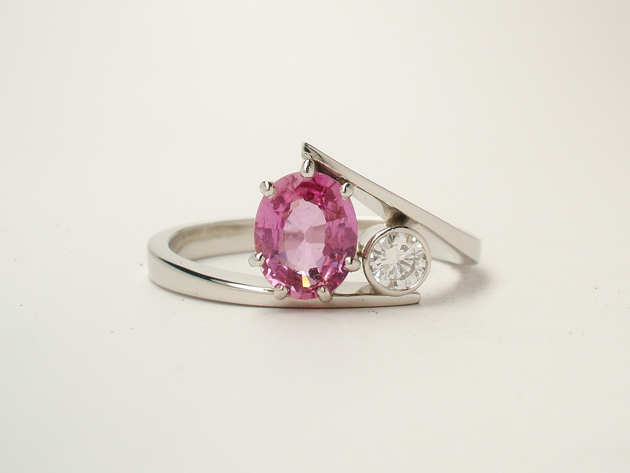 A 2 stone oval peg set pink sapphire and rub-over set round brilliant cut diamond straight wishbone cross-over ring mounted in platinum.