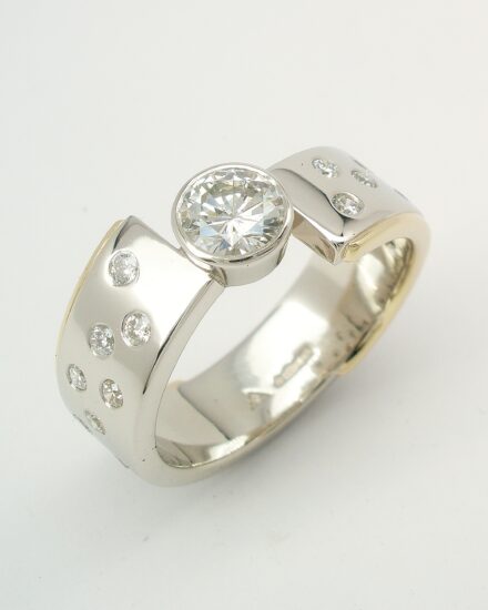 A single stone rub-over set round brilliant cut diamond ring with broad flat sectioned cross-over style band flush set with small round diamonds and edged on diagonally opposite sides with 9ct. gold.