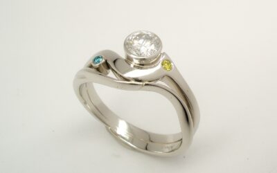 A rub-over set single stone round brilliant cut diamond 'Wave' style ring with a small rub-over set round sky blue diamond and a flush set canary yellow diamond.