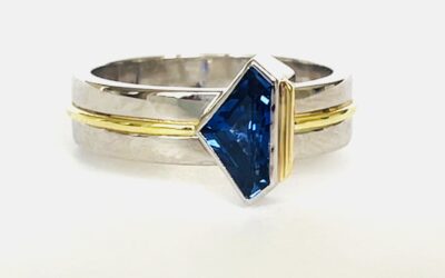 A single stone 'kite' shaped sapphire ring rub-over set in platinum with an 18ct yellow gold wire applied along the back edge and mounted on a 6mm broad platinum band with a part inlayed 18ct green gold round wire around the centre.