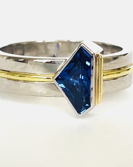 A single stone 'kite' shaped sapphire ring rub-over set in platinum with an 18ct yellow gold wire applied along the back edge and mounted on a 6mm broad platinum band with a part inlayed 18ct green gold round wire around the centre.