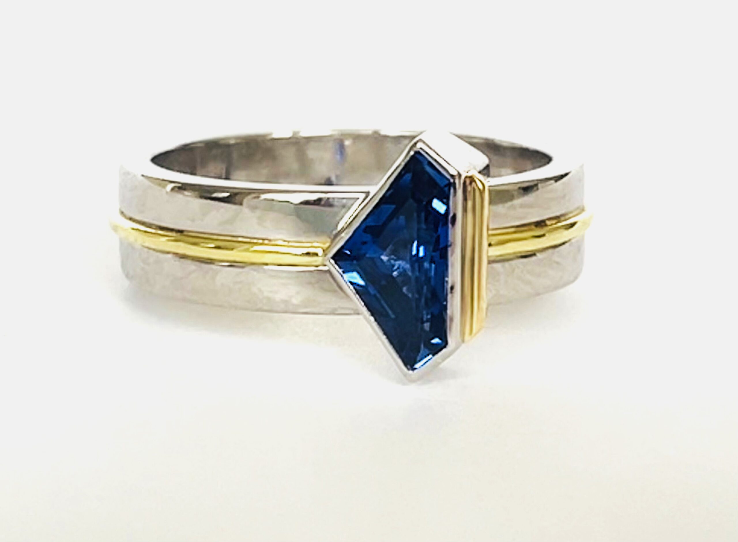A single stone 'kite' shaped sapphire ring rub-over set in platinum with an 18ct yellow gold wire applied along the back edge and mounted on a 6mm broad platinum band with a part inlayed 18ct green gold round wire around the centre.