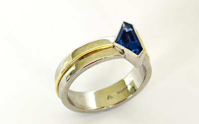 A single stone 'kite' shaped sapphire ring rub-over set in platinum with an 18ct yellow gold wire applied along the back edge and mounted on a 6mm broad platinum band with a part inlayed 18ct green gold round wire around the centre.