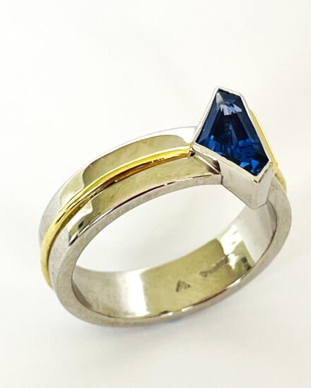 A single stone 'kite' shaped sapphire ring rub-over set in platinum with an 18ct yellow gold wire applied along the back edge and mounted on a 6mm broad platinum band with a part inlayed 18ct green gold round wire around the centre.