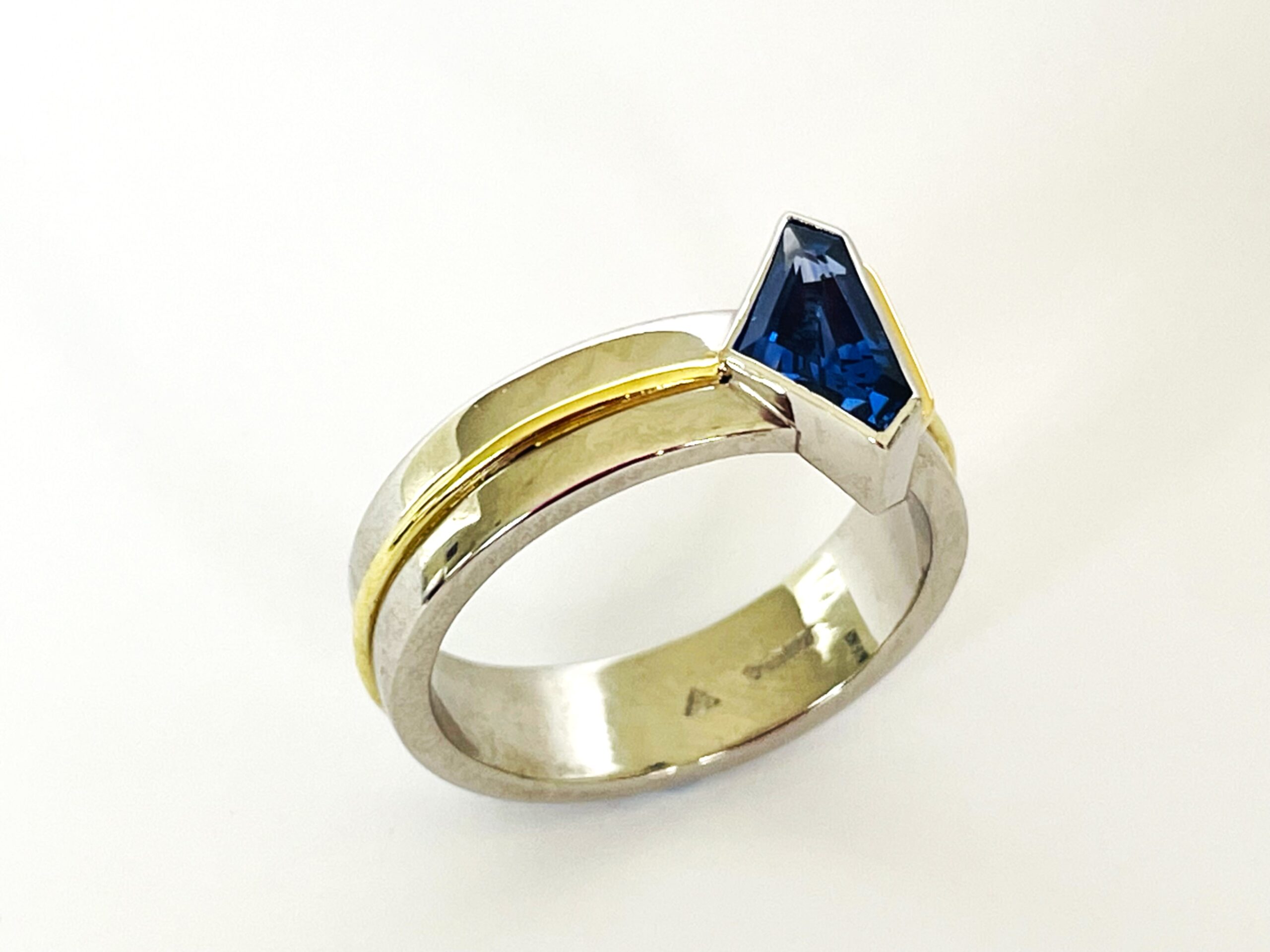 A single stone 'kite' shaped sapphire ring rub-over set in platinum with an 18ct yellow gold wire applied along the back edge and mounted on a 6mm broad platinum band with a part inlayed 18ct green gold round wire around the centre.