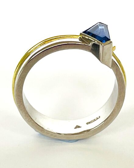 A single stone 'kite' shaped sapphire ring rub-over set in platinum with an 18ct yellow gold wire applied along the back edge and mounted on a 6mm broad platinum band with a part inlayed 18ct green gold round wire around the centre.