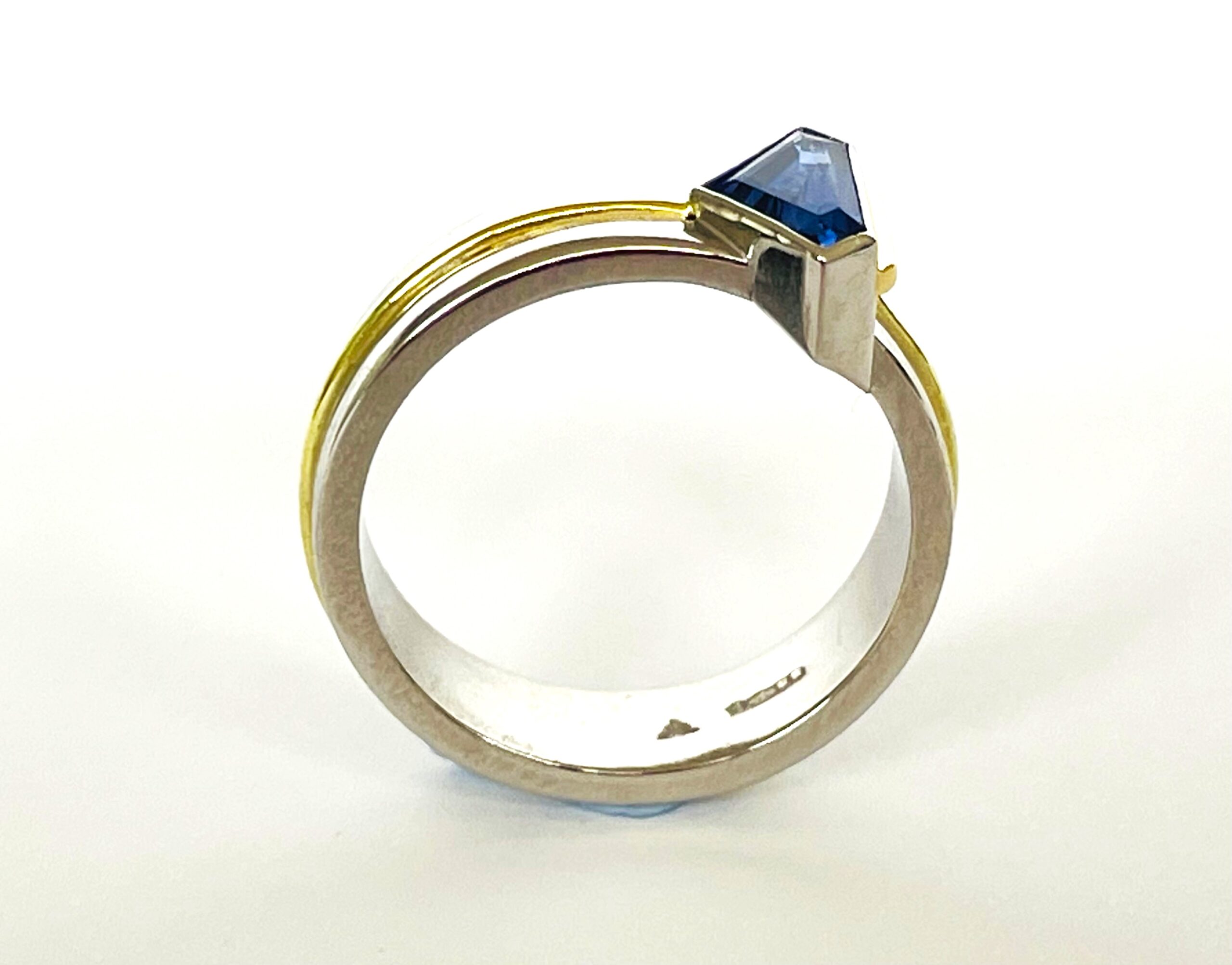 A single stone 'kite' shaped sapphire ring rub-over set in platinum with an 18ct yellow gold wire applied along the back edge and mounted on a 6mm broad platinum band with a part inlayed 18ct green gold round wire around the centre.